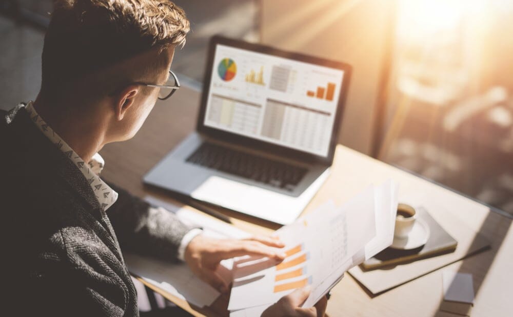 a man looking through papers with charts on them while working on his laptop