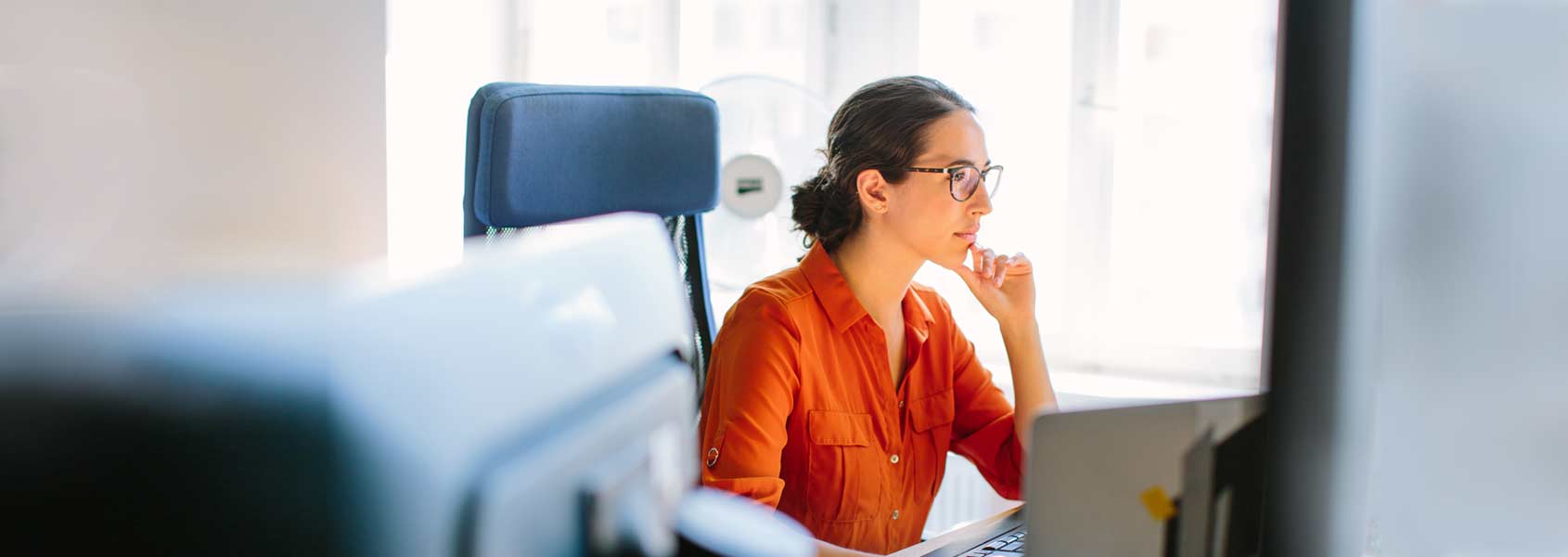 A women in a red blouse and glasses sitting by a window concentrating on a computer screen