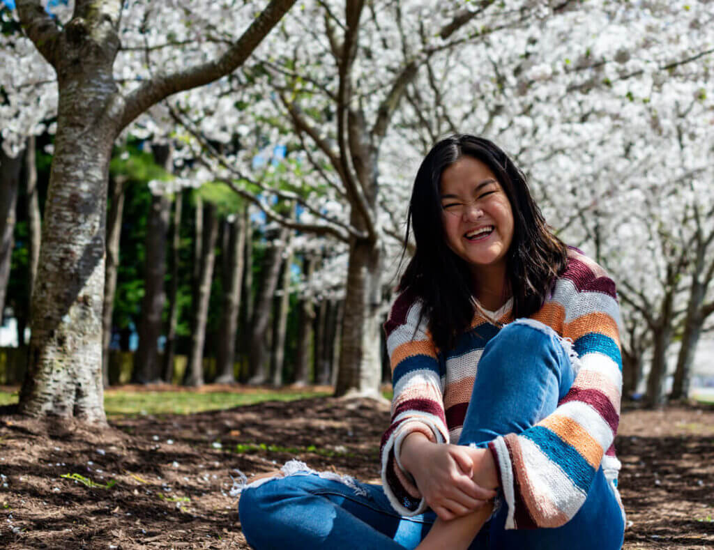 A woman smiling and sitting on the ground beneath blossoming trees